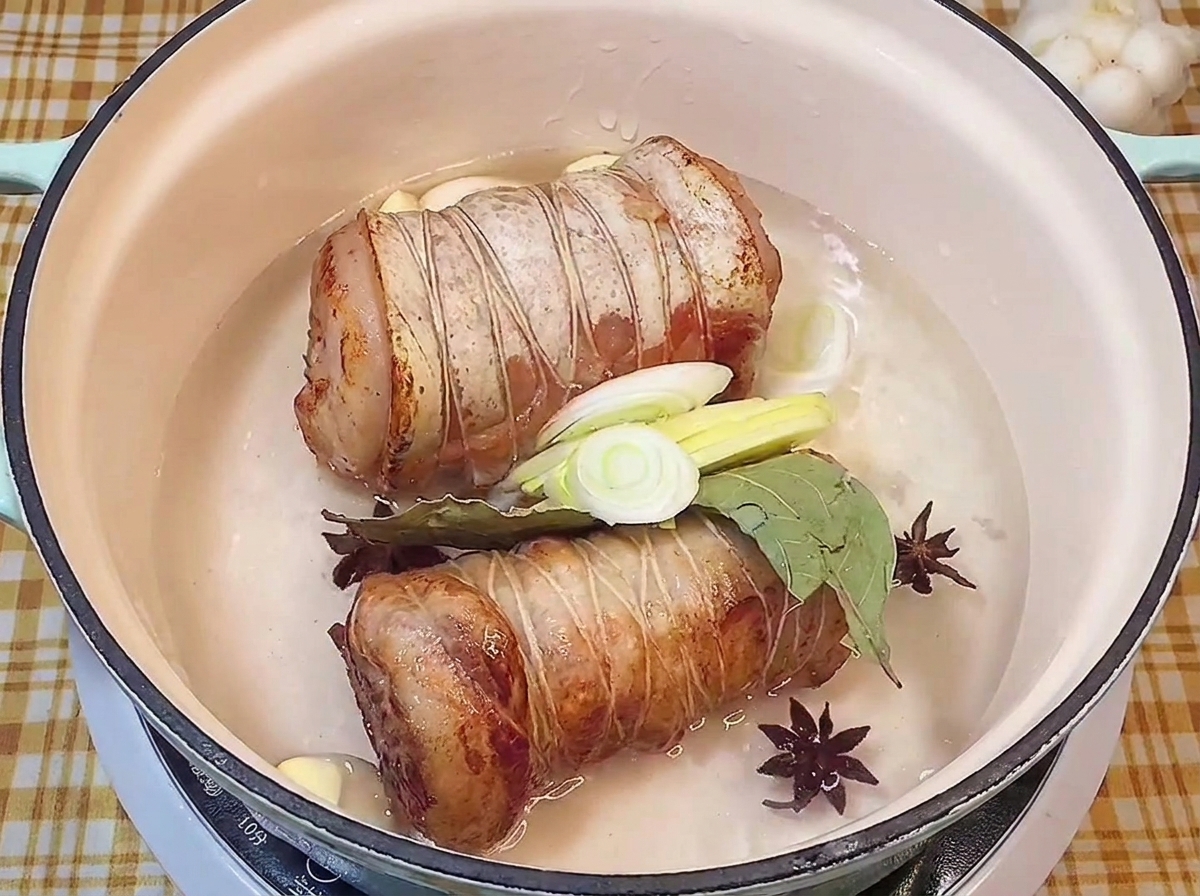 Rollos de cerdo en una olla con líquido, anís estrellado, laurel, ajo y puerro.