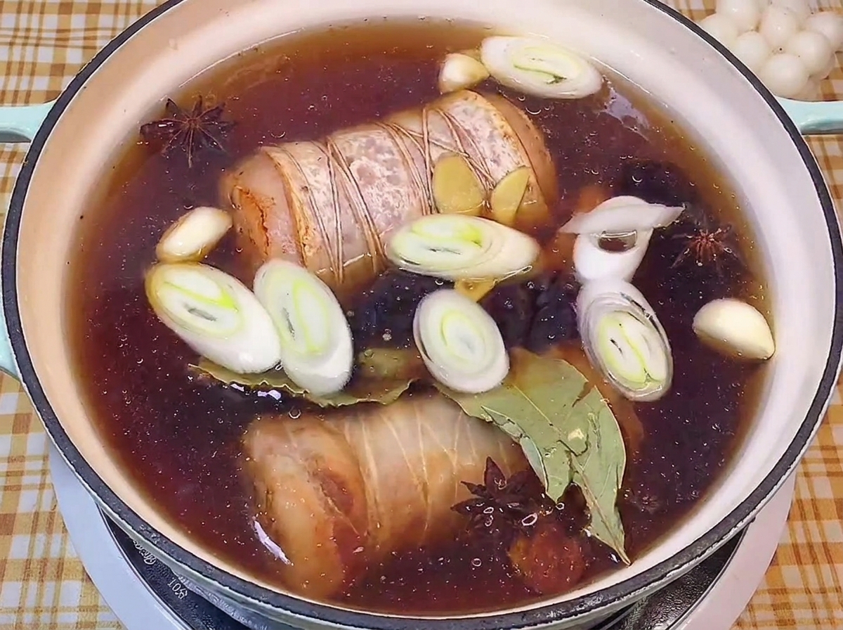 Panceta enrollada cociéndose en un caldo oscuro con puerros, ajo y anís estrellado.