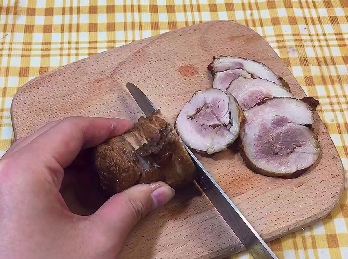 Mano usando un cuchillo para cortar el rollo de chashu en rodajas finas sobre una tabla de madera.