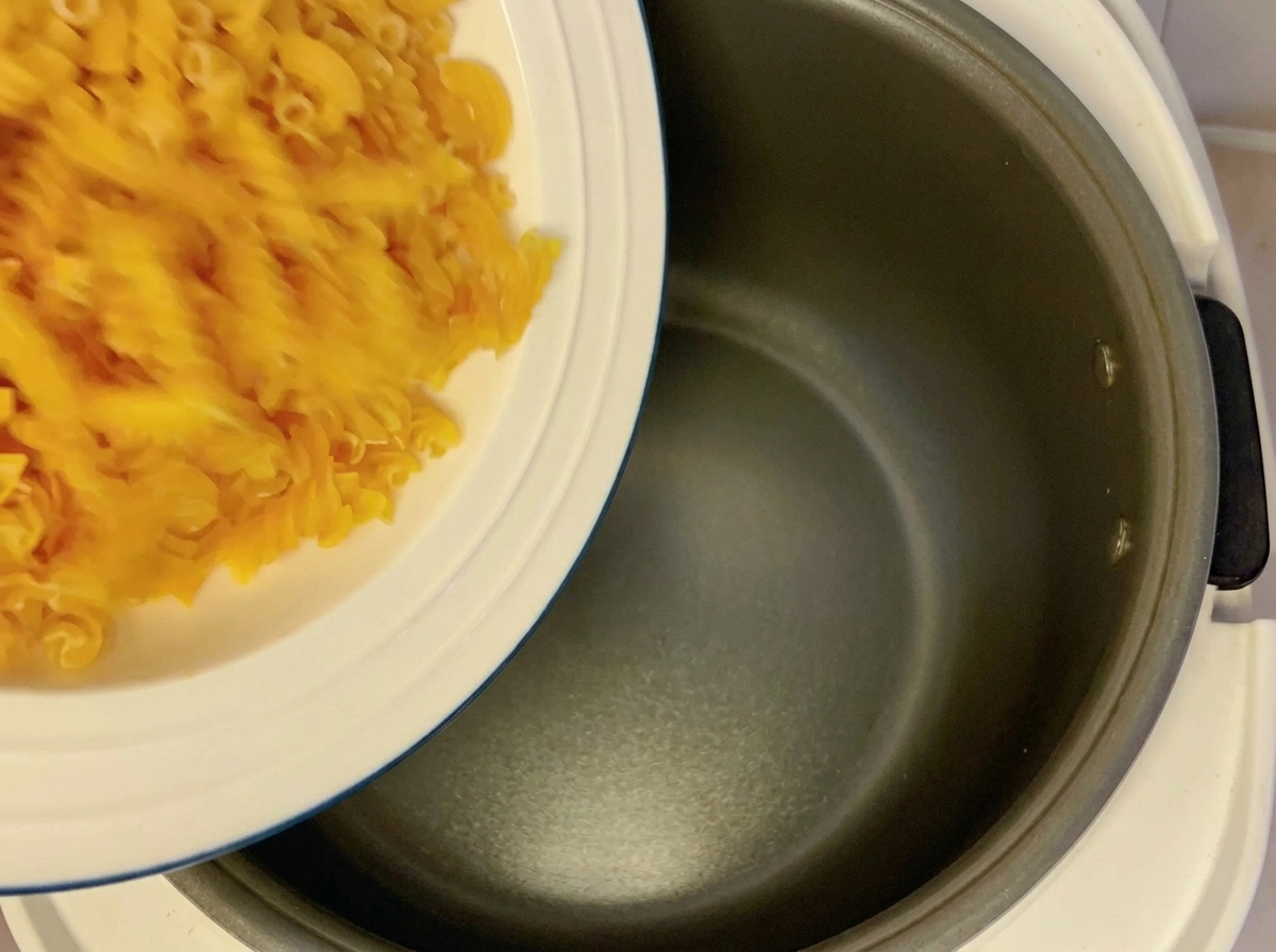 Pasta fusilli seca vertiéndose desde un plato blanco en el recipiente oscuro y vacío de una arrocera.