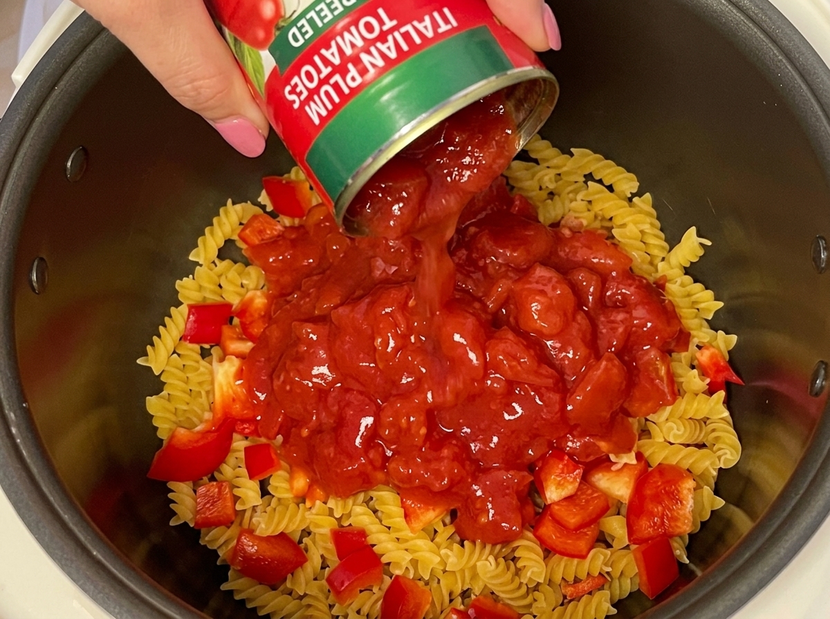 Una mano vertiendo una lata de tomates pelados troceados sobre la pasta y el pimiento rojo.