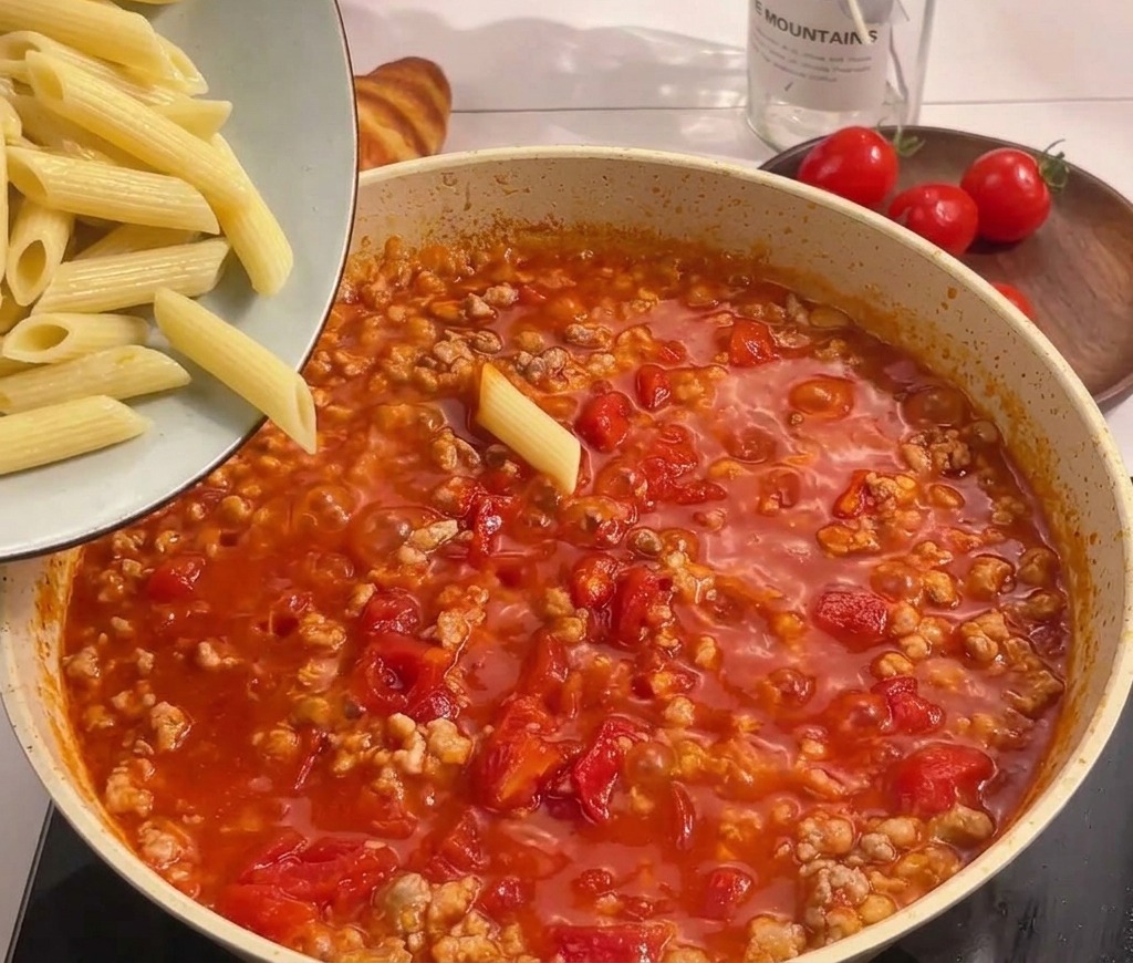 Pasta penne hervida vertiéndose desde un bol a una sartén con salsa de tomate y carne hirviendo.