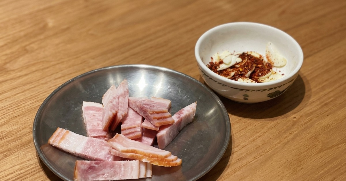 Bacon crudo en láminas junto a un pequeño cuenco con ajo laminado y copos de chile sobre una mesa de madera.