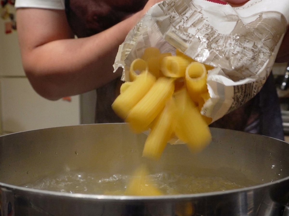 Pasta rigatoni seca cayendo de una bolsa de papel a una olla con agua hirviendo a borbotones.