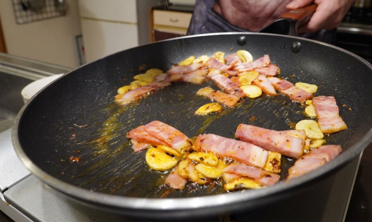 Bacon, ajo y copos de chile cocinándose en una sartén mientras se sazonan con pimienta negra recién molida.