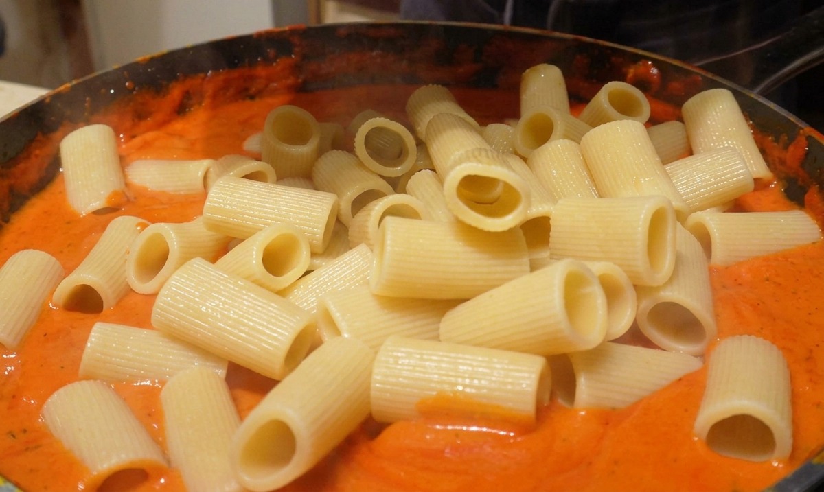 Añadiendo rigatoni cocido a una sartén con una salsa suave de color naranja rojizo.