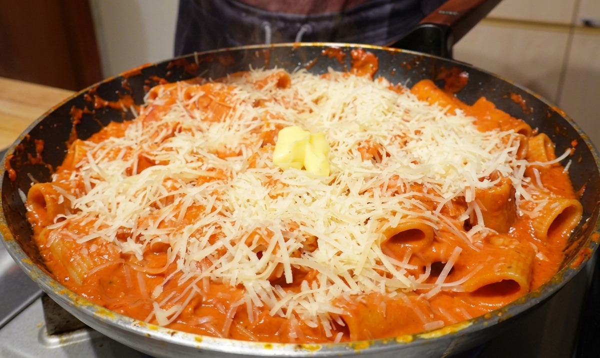 Un dado de mantequilla amarilla sobre una montaña de queso parmesano y pasta rigatoni en la sartén caliente.