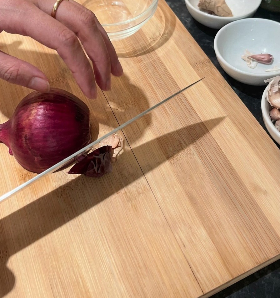Una mano sujetando una cebolla roja sobre una tabla de cortar de madera con un cuchillo de chef listo para picar.
