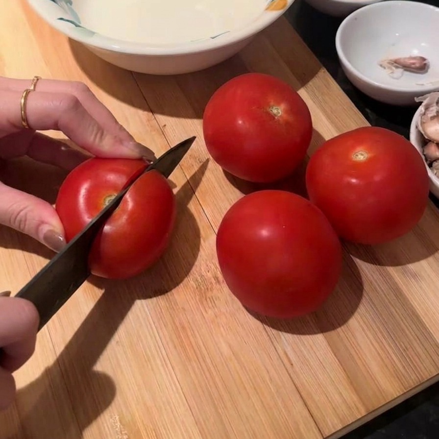 Manos usando un cuchillo de chef para cortar un tomate rojo fresco sobre una tabla de madera con más tomates cerca.