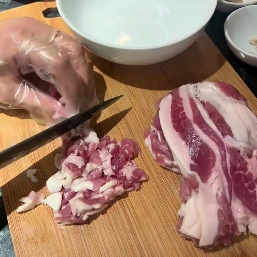 Una mano con guante usando un cuchillo negro para cortar tiras de beicon crudo en trozos pequeños sobre una tabla de madera.