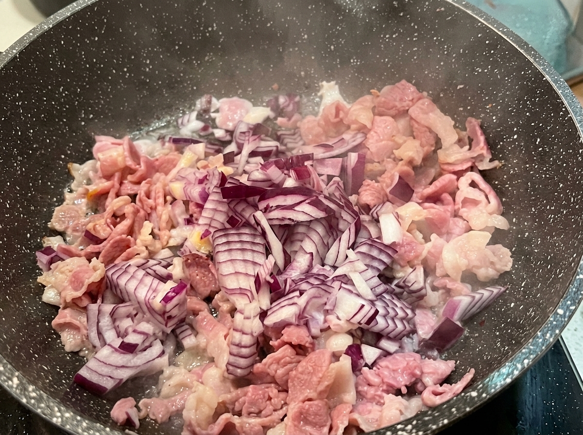 Cebolla roja picada esparcida sobre trozos de beicon parcialmente cocinados en una sartén moteada caliente.