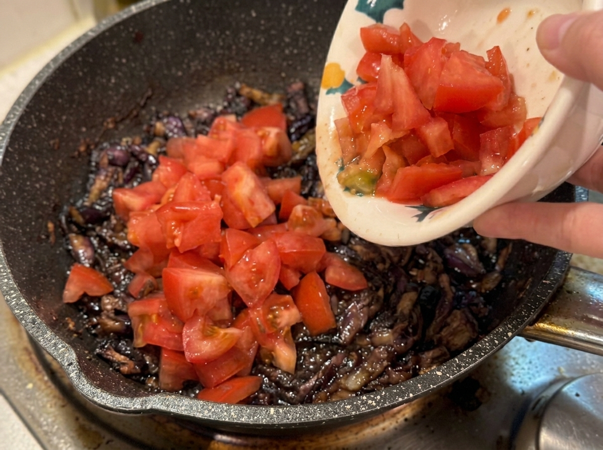 Una mano sosteniendo un cuenco de cerámica para verter tomates frescos picados en una sartén con beicon y cebolla.