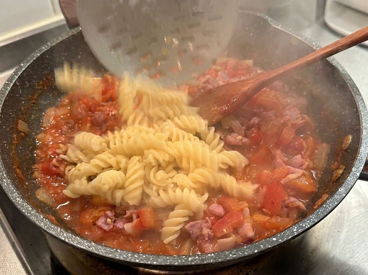 Pasta fusilli cocida cayendo desde una espumadera a una sartén caliente con salsa roja de tomate y beicon.