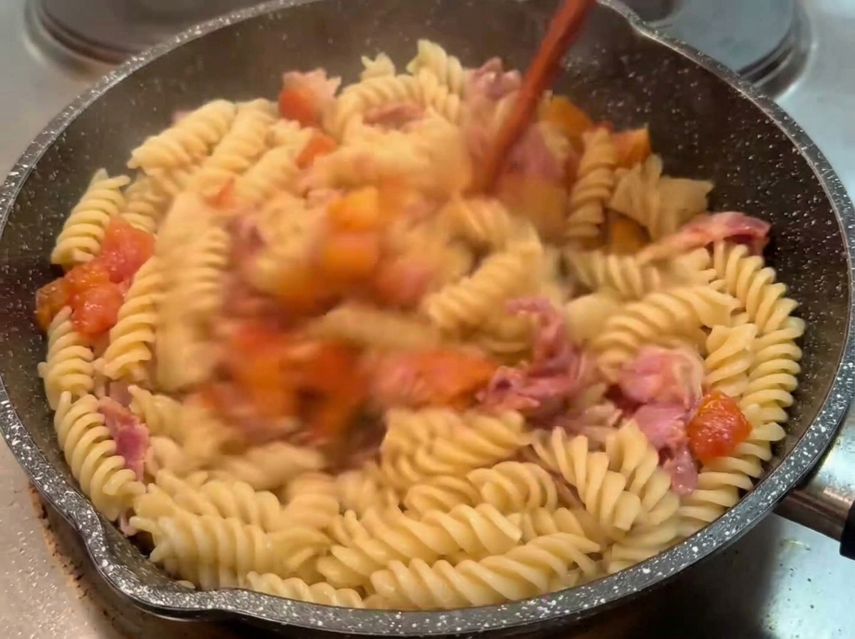 Pasta fusilli cocida mezclándose en una sartén oscura con salsa de tomate y beicon usando un utensilio de madera.