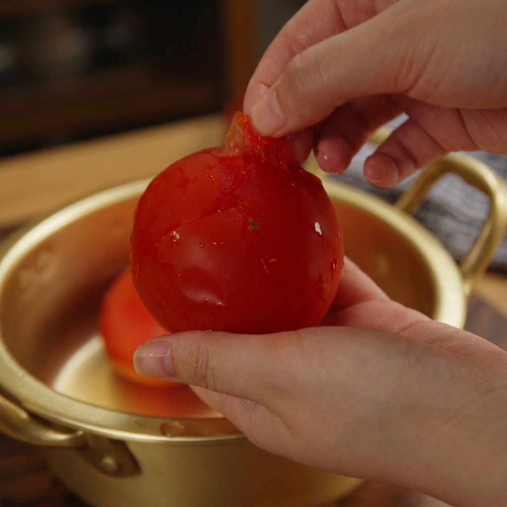 Manos retirando la piel de un tomate rojo brillante sobre un cuenco.