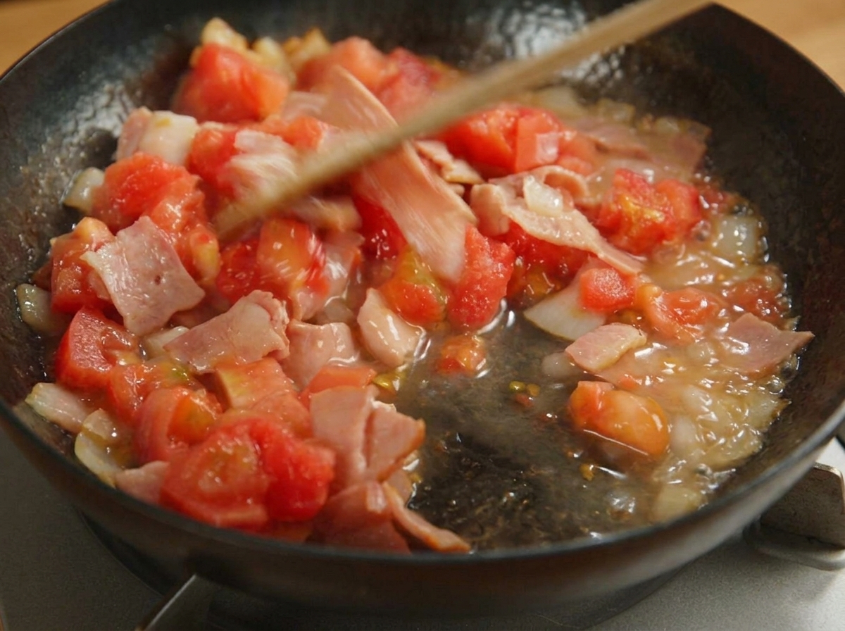 Espátula removiendo tomates picados, bacón y cebolla en una sartén negra.