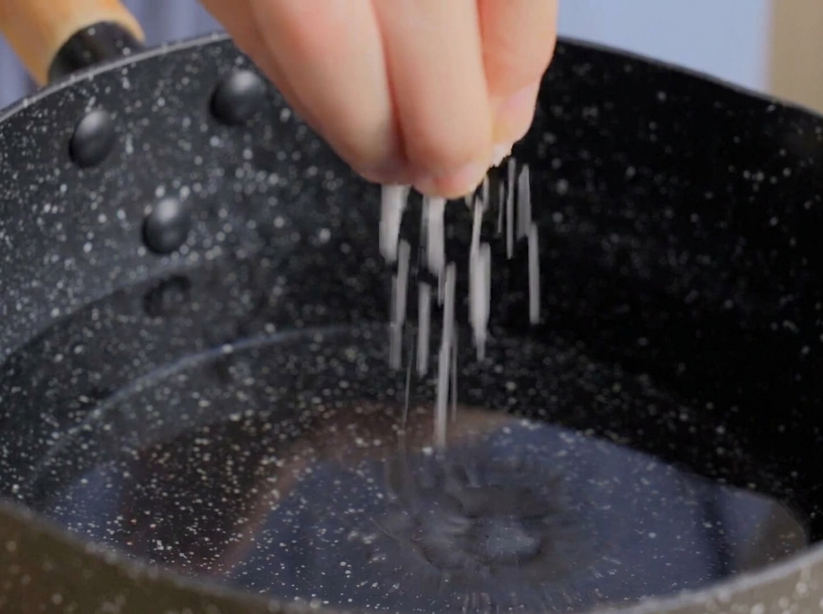 Mano espolvoreando una pizca de sal gruesa en una olla oscura con agua.
