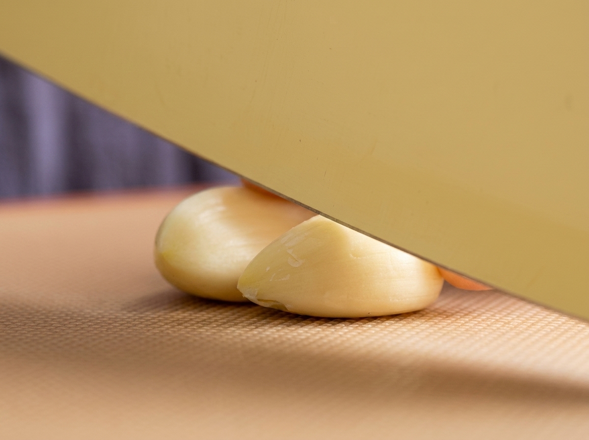 Un cuchillo de cocina ancho descansando sobre dos dientes de ajo pelados en una tabla de cortar de textura marrón, listos para ser picados.
