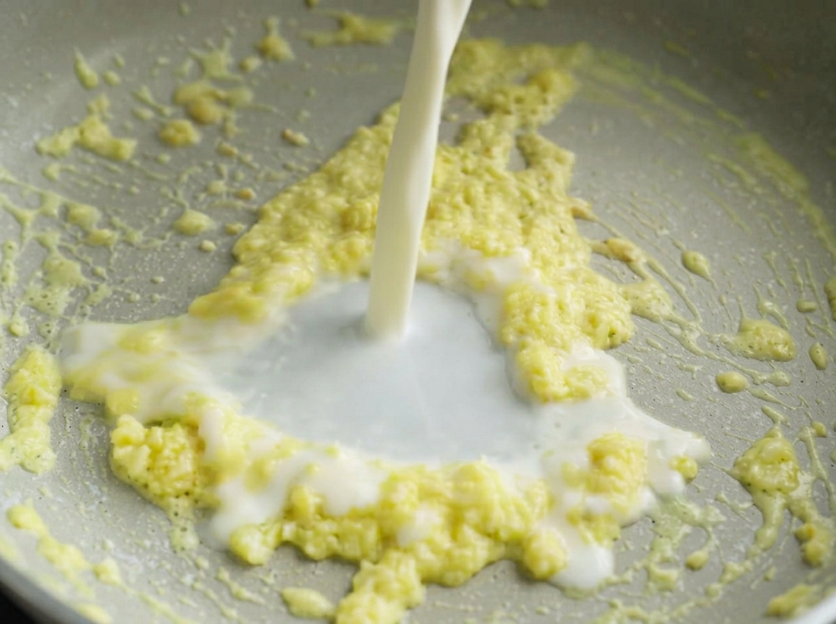 Un chorrito de leche blanca vertiéndose en una sartén que contiene una mezcla chisporroteante de mantequilla y ajo picado.