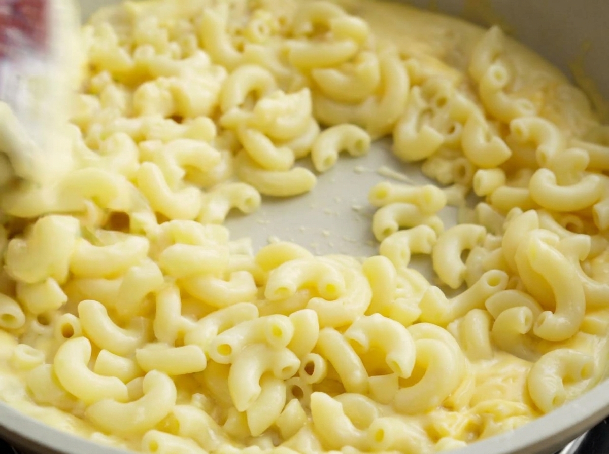 Una espátula revolviendo macarrones cocidos en una sartén clara, cubriendo completamente la pasta con cremosa salsa de queso.