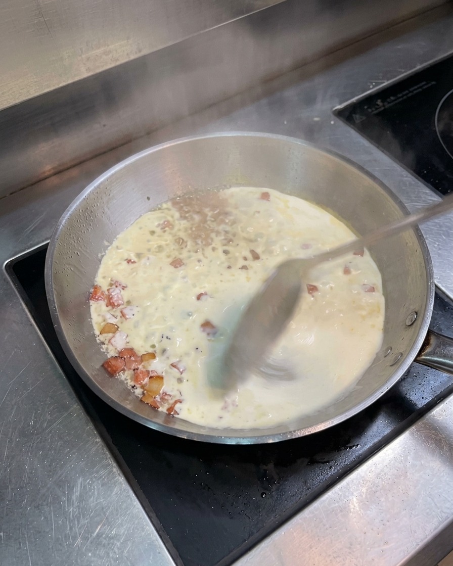 Una sartén de acero inoxidable con nata y bacón hirviendo a fuego lento, removida con una cuchara metálica.