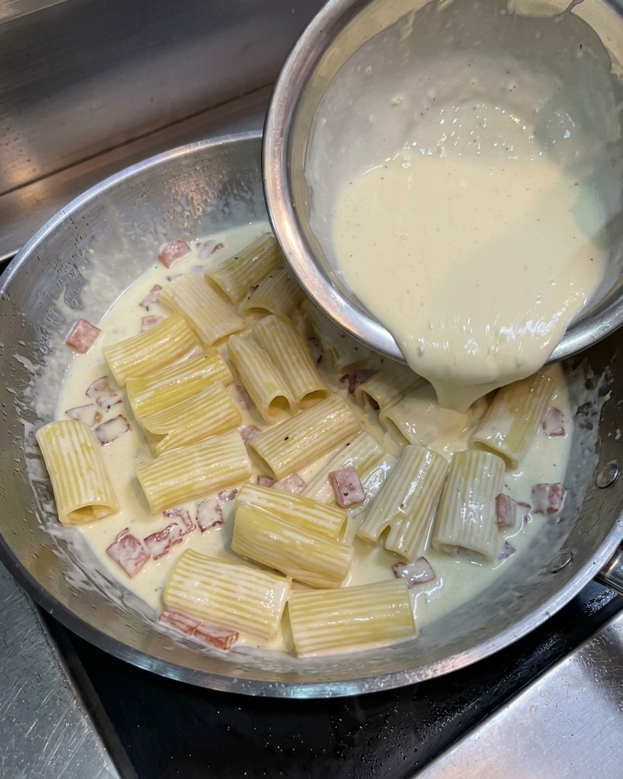 Vertiendo una mezcla amarilla de yema, nata y queso desde un cuenco sobre los rigatoni y el bacón en la sartén.