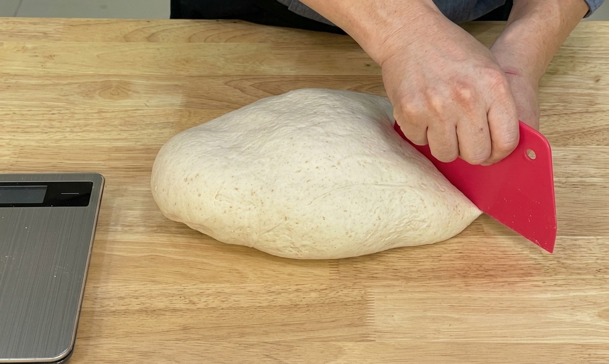 Un chef usando una rasqueta roja de panadero para dividir una pieza grande de masa de pizza lisa sobre una superficie de madera.