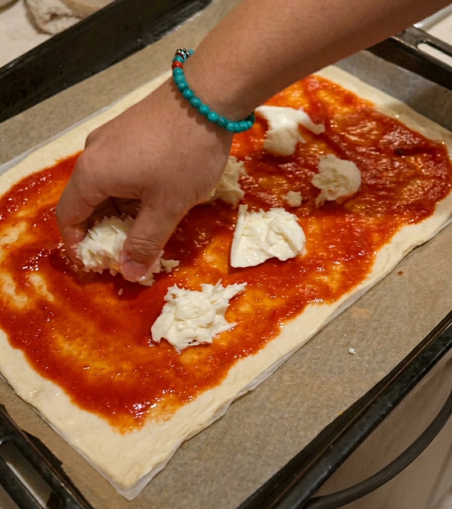 Una mano colocando trozos irregulares de queso mozzarella blanco sobre una masa de pizza cubierta con salsa roja.