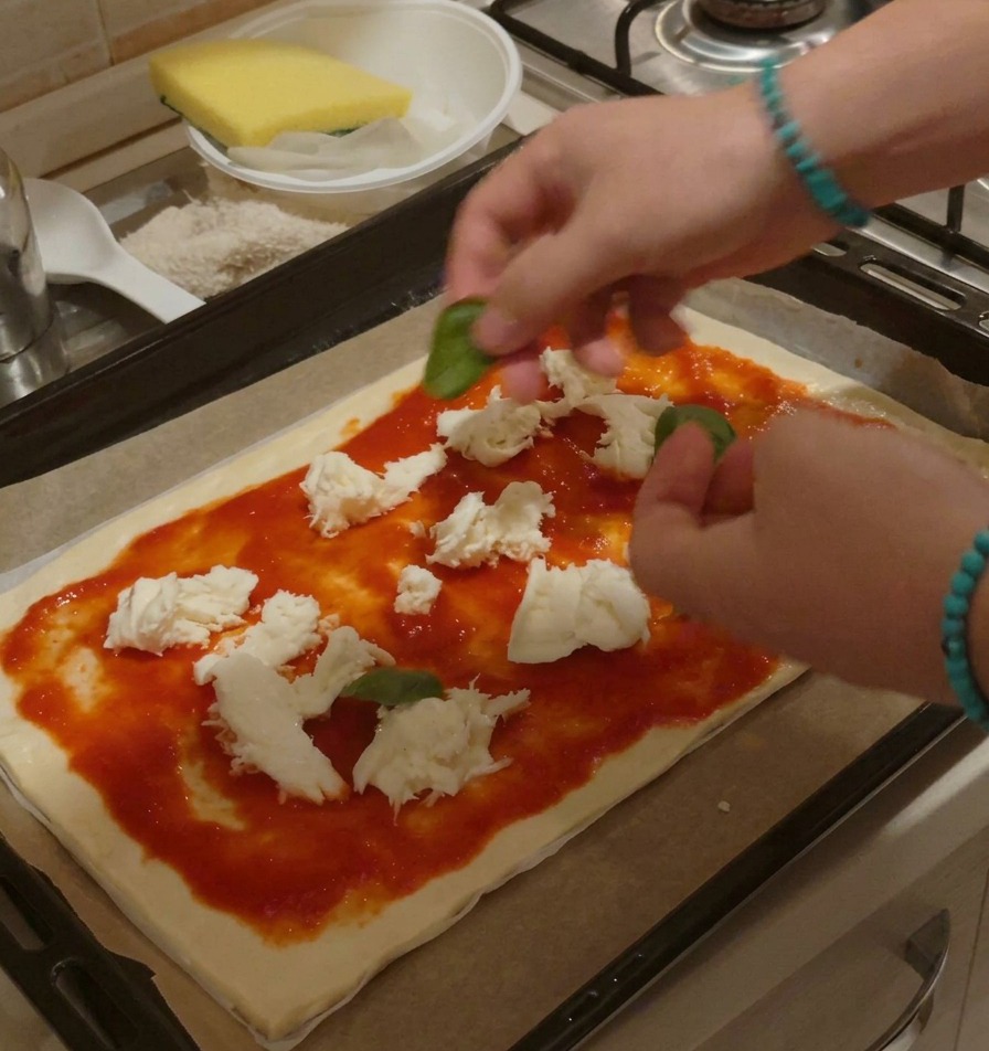 Manos colocando cuidadosamente hojas enteras de albahaca verde sobre una pizza cruda con tomate y mozzarella.