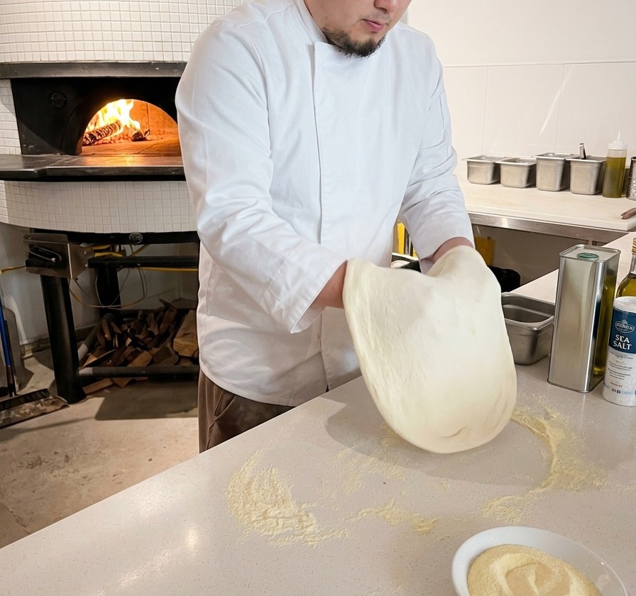 Un chef presionando y estirando una masa redonda de pizza sobre una encimera blanca espolvoreada con harina.