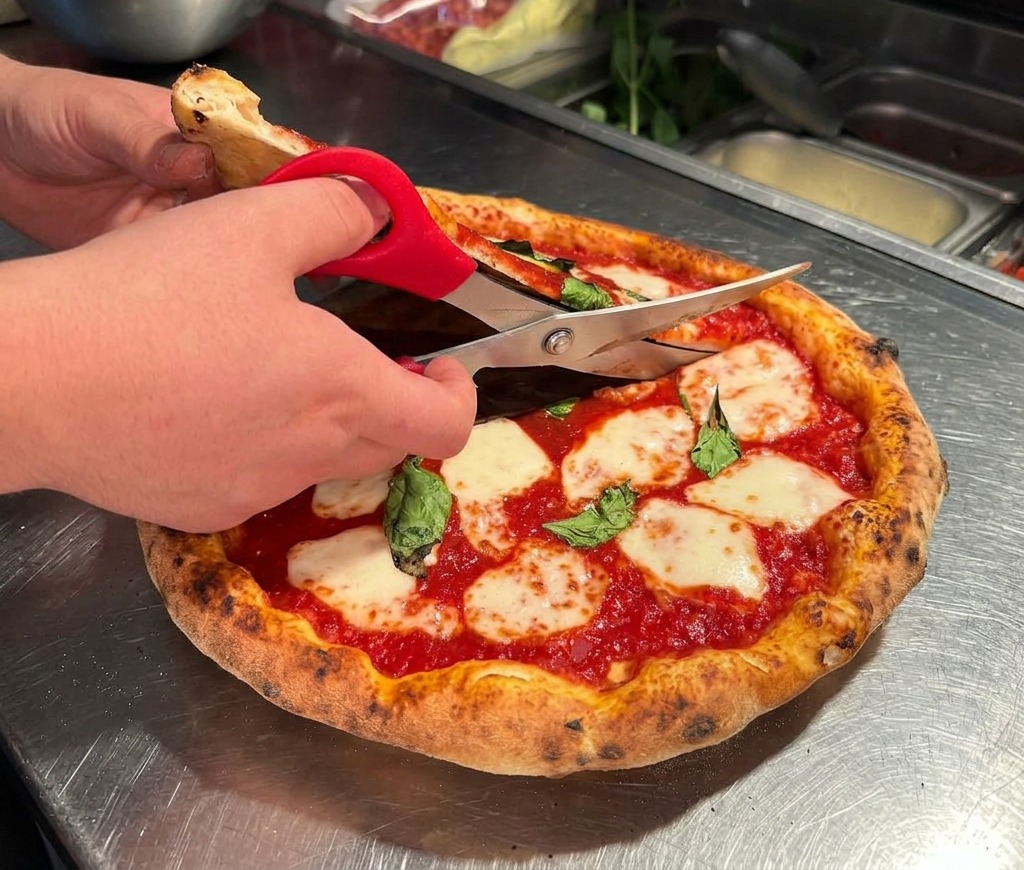 Manos usando tijeras de cocina de mango rojo para cortar una pizza Margarita caliente sobre una encimera de acero inoxidable.