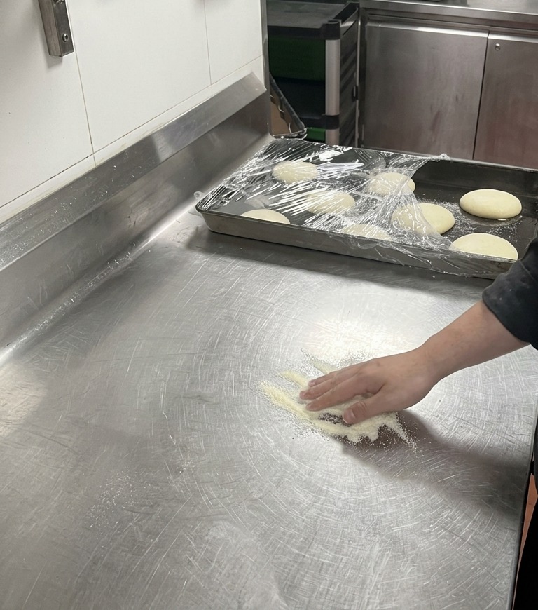 Mano de un chef presionando suavemente una bola de masa de pizza sobre un montón de harina blanca en una encimera de metal.