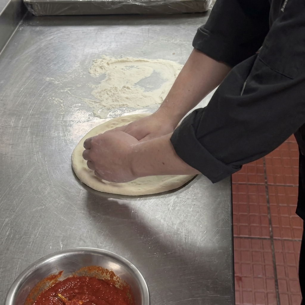 Manos presionando el centro de una masa de pizza aplanada, dejando un borde grueso alrededor.