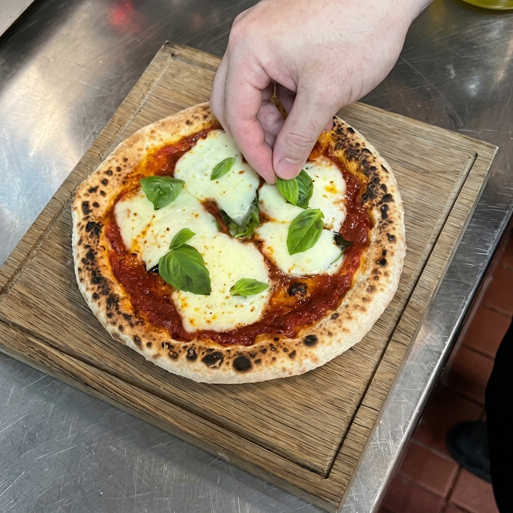 Una mano colocando hojas de albahaca verde sobre una pizza caliente con bordes crujientes apoyada en una tabla de madera.