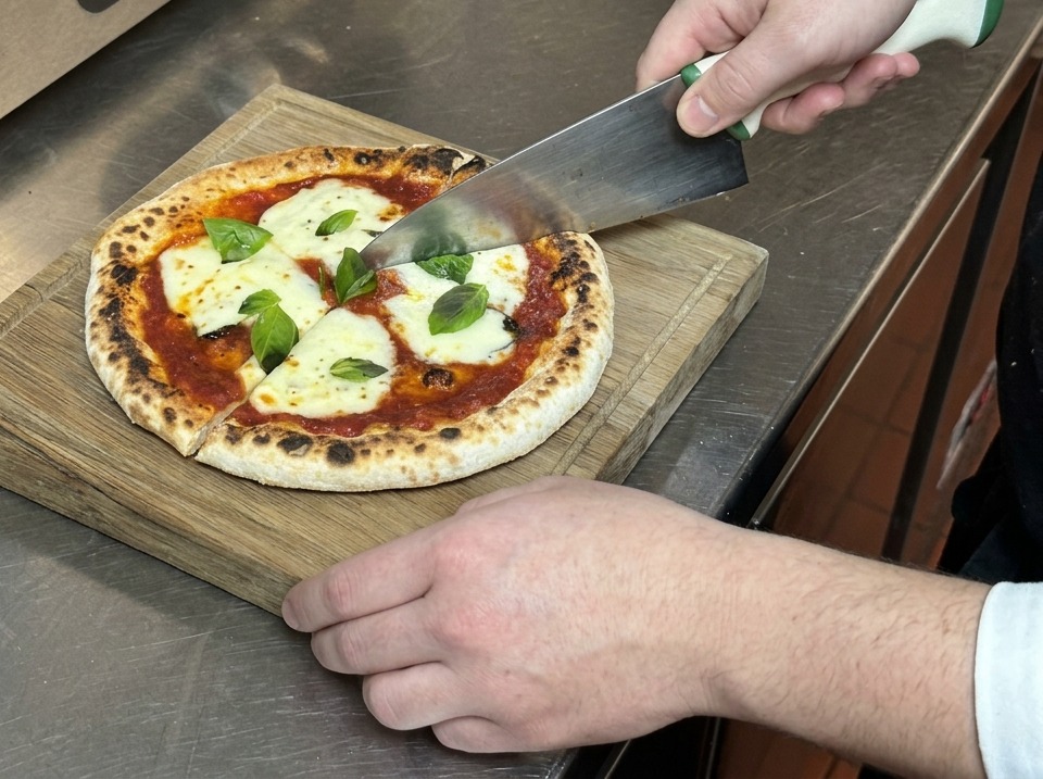 Manos usando un cuchillo grande para cortar una pizza Margarita fresca en porciones sobre una tabla de madera.