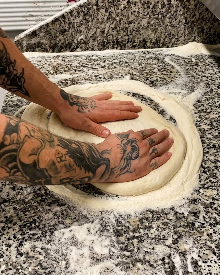 Manos tatuadas presionando y estirando suavemente una pieza redonda de masa de pizza sobre una superficie de mármol enharinada.