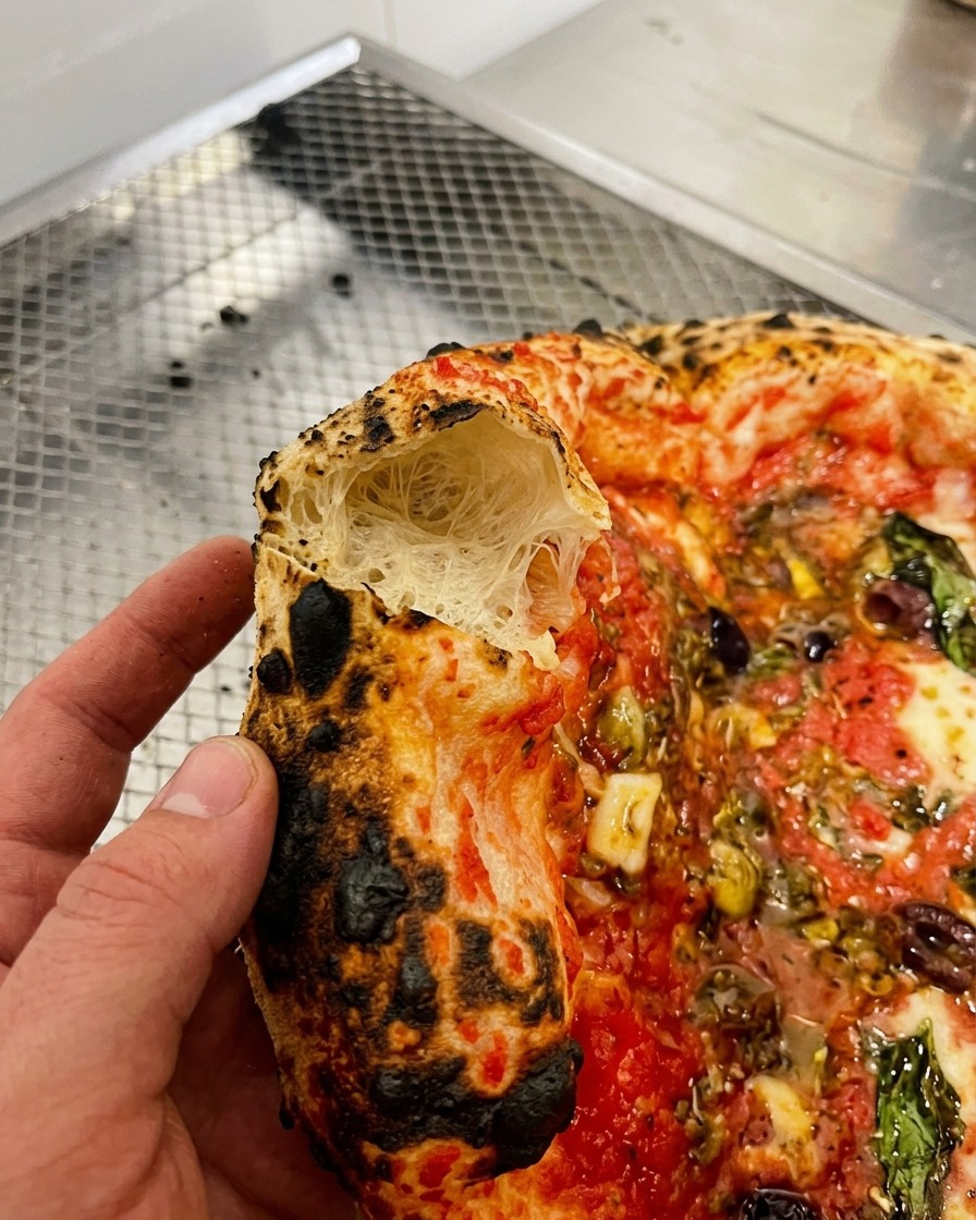 Primer plano de una mano sosteniendo un trozo de borde de pizza abierto mostrando una miga interior aireada y alveolada.