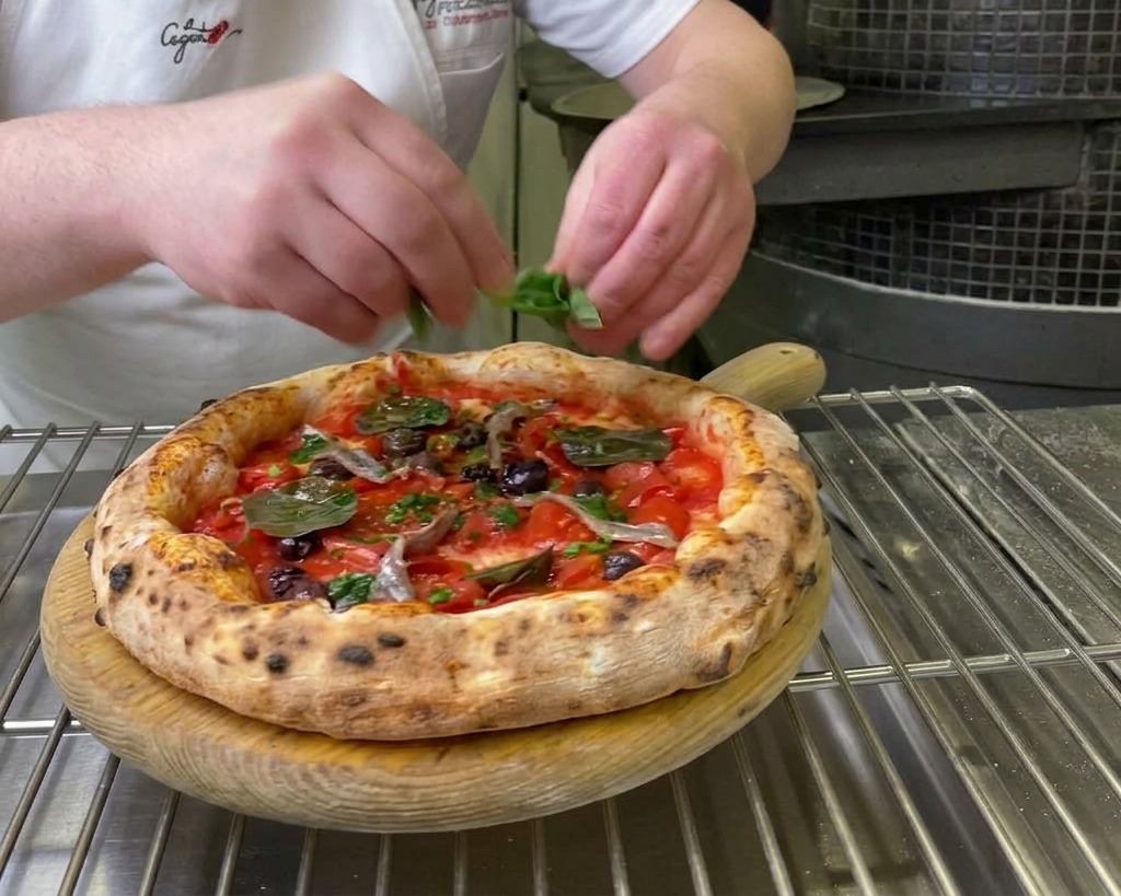 Manos de chef colocando hojas verdes de albahaca fresca sobre una pizza Marinara recién horneada.