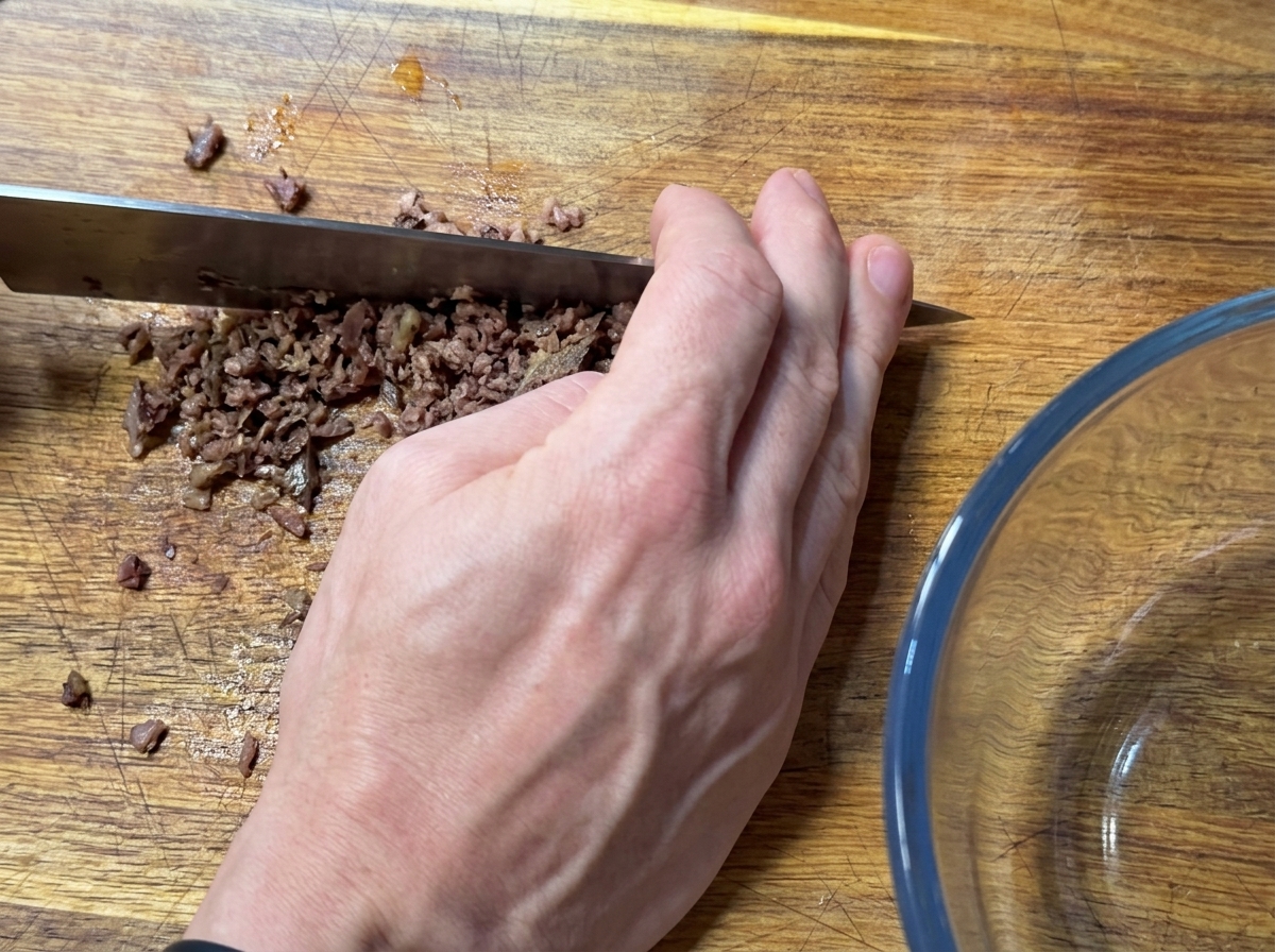 Una mano usando un cuchillo grande para picar finamente filetes de anchoa sobre una tabla de madera.