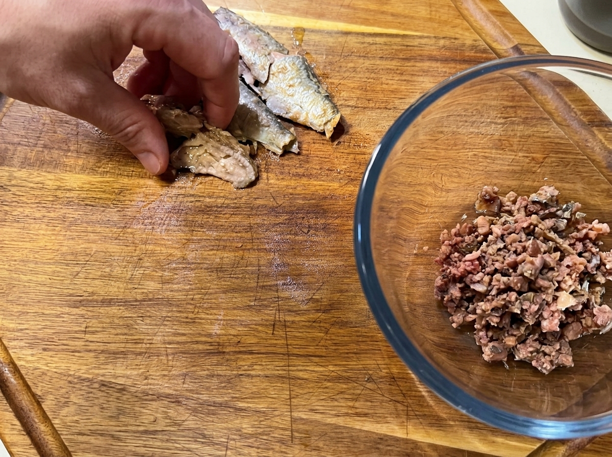 Una mano desmenuzando filetes de sardina sin espinas sobre una tabla junto a un bol con pescado picado.