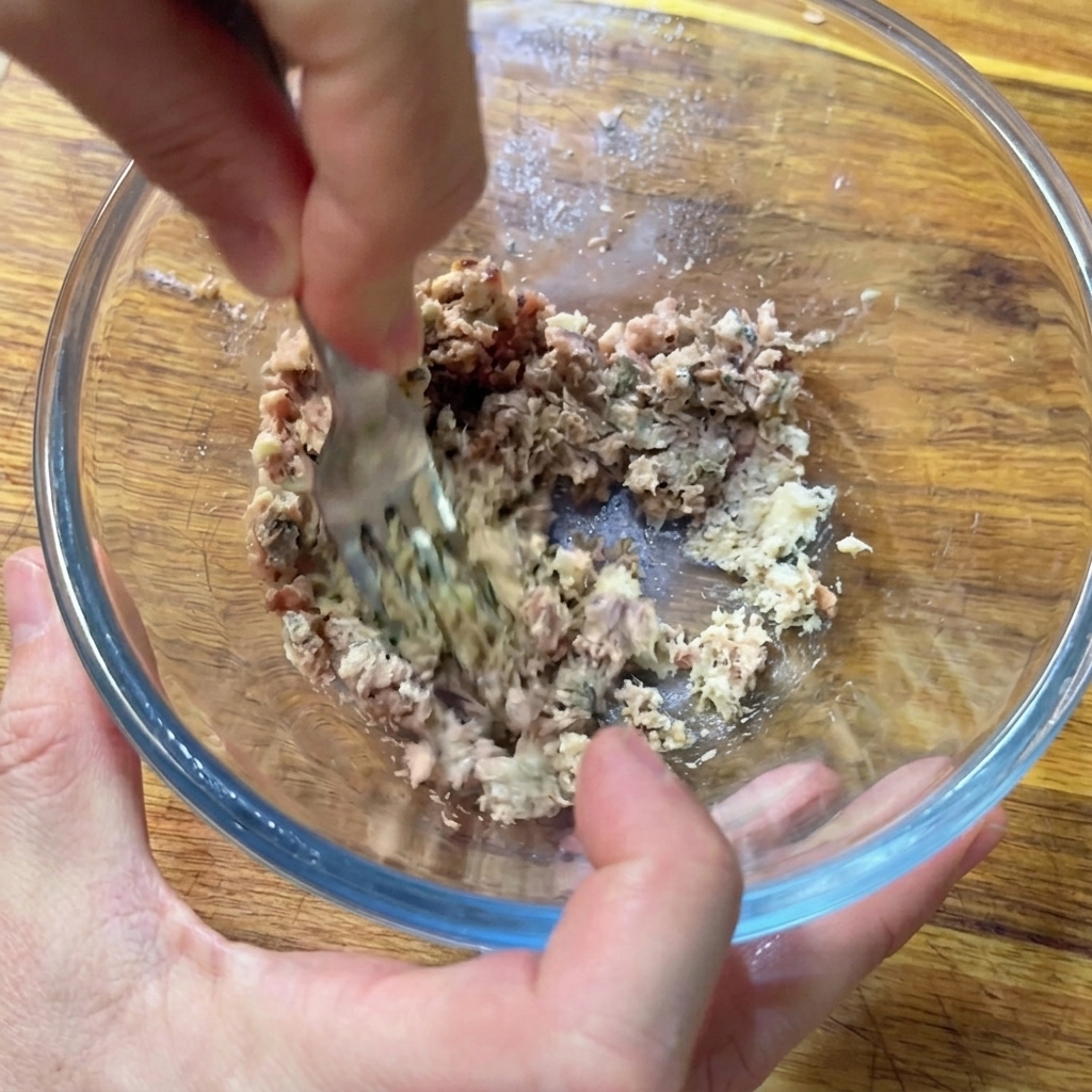 Manos usando un tenedor de metal para machacar anchoas y sardinas picadas en un bol de cristal hasta crear una pasta.