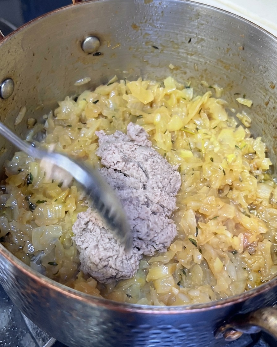 Una cucharada de pasta de pescado grisácea siendo añadida a una olla de cebollas profundamente caramelizadas.