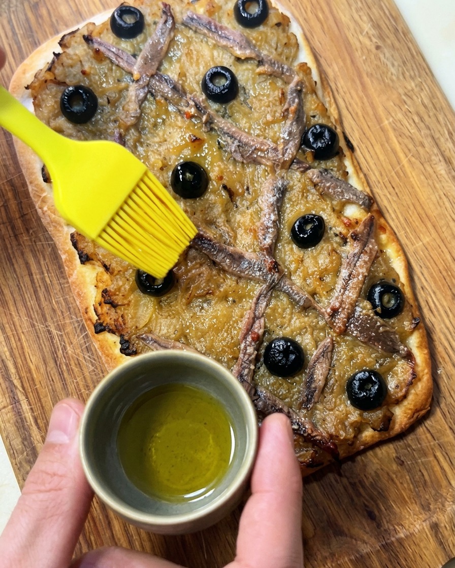 Una mano usando un pincel de silicona para aplicar aceite de oliva sobre una Pissaladière recién horneada.