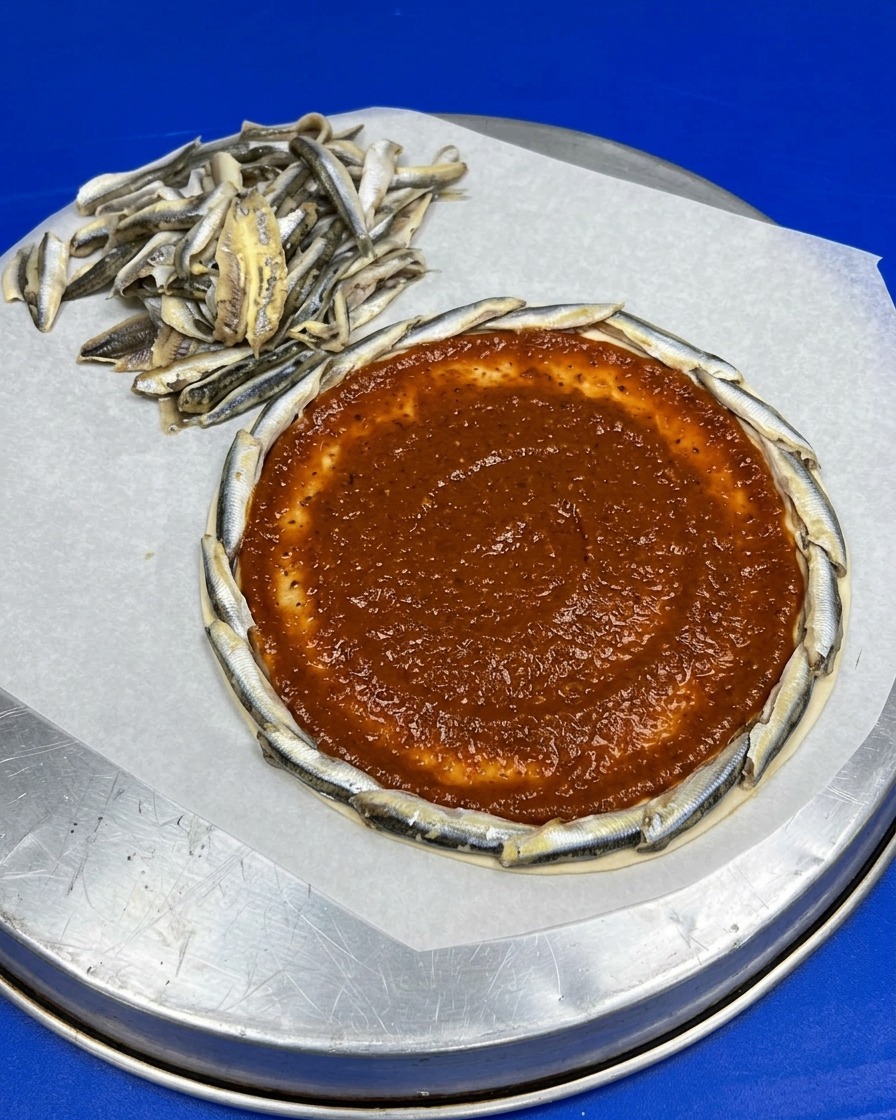 Base de pizza redonda con un borde de boquerones y una capa de salsa de tomate en el centro.