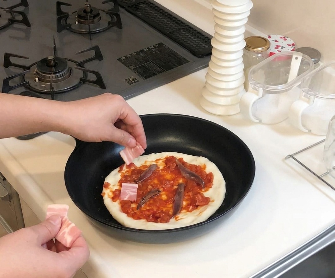 Manos colocando pequeños trozos de tocino crudo sobre una masa de pizza cubierta con salsa roja y anchoas en una sartén.