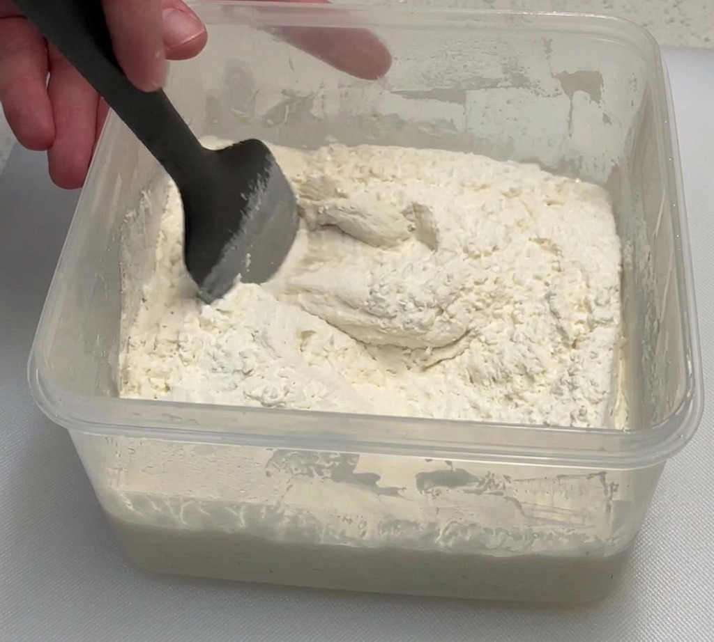 Una mano usando una espátula negra para mezclar harina y poolish en un recipiente transparente.