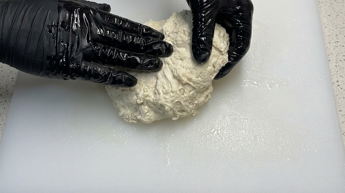 Manos con guantes negros amasando una masa de pizza húmeda sobre una tabla blanca.