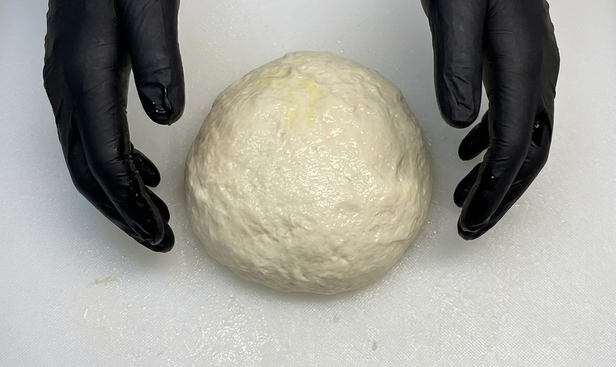Manos con guantes dando forma a una bola de masa de pizza suave y redonda sobre una tabla blanca.