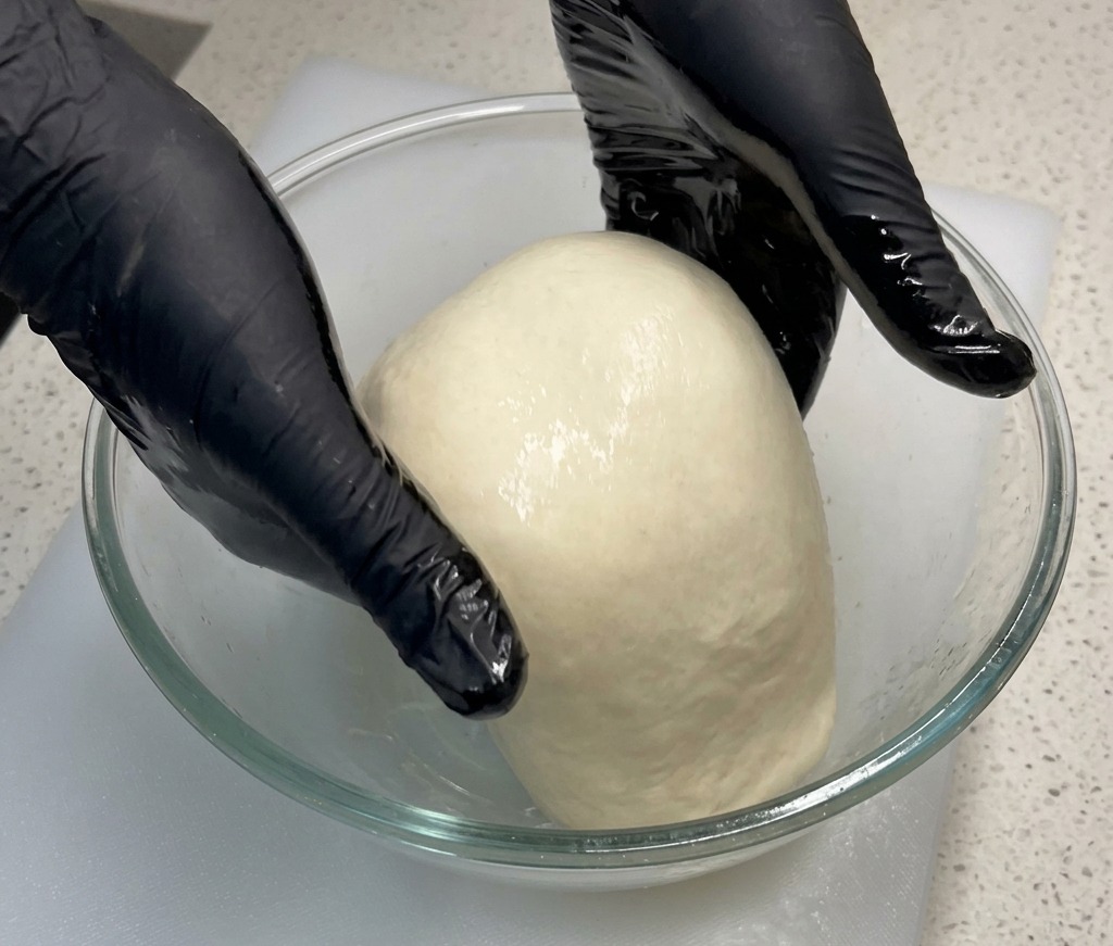 Manos levantando y estirando el centro de una masa de pizza hacia arriba dentro de un bol de cristal.