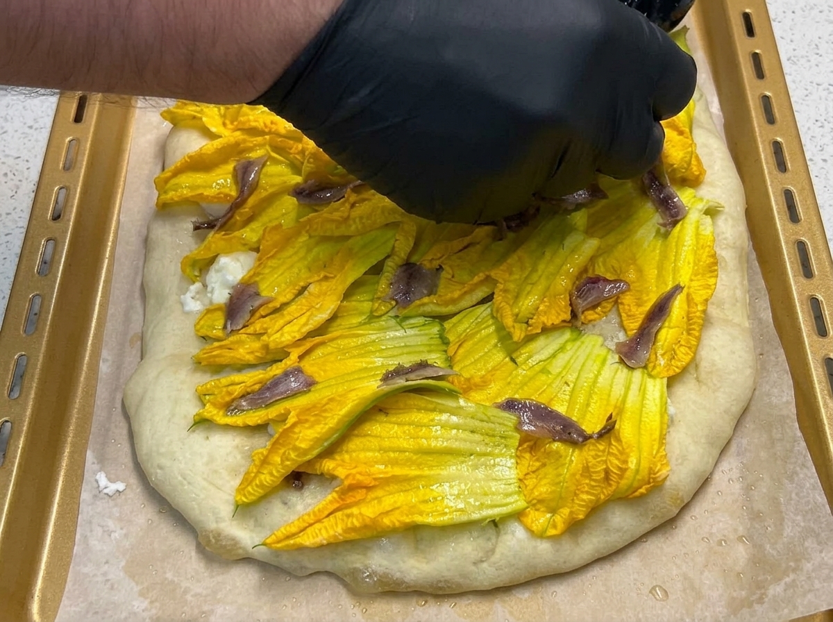 Mano colocando filetes de anchoa sobre las flores de calabaza en la pizza.